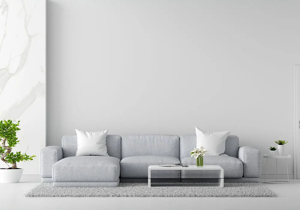 یک مبل ال بزرگ توسی رنگ با دو کوسن سفید که روی یک فرش ساده توسی و در یک اتاق توسی و سفید است