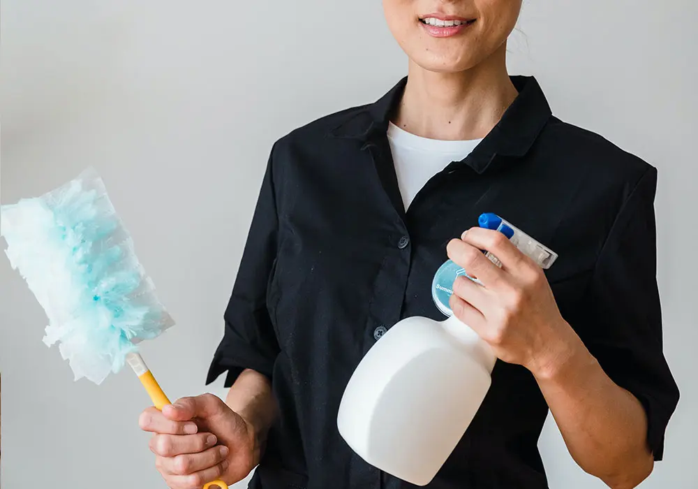 تصویر نیمه ای از یک خانم که در دستانش اسپری تمیز کننده و یک وسیله برای گردگیری وجود دارد