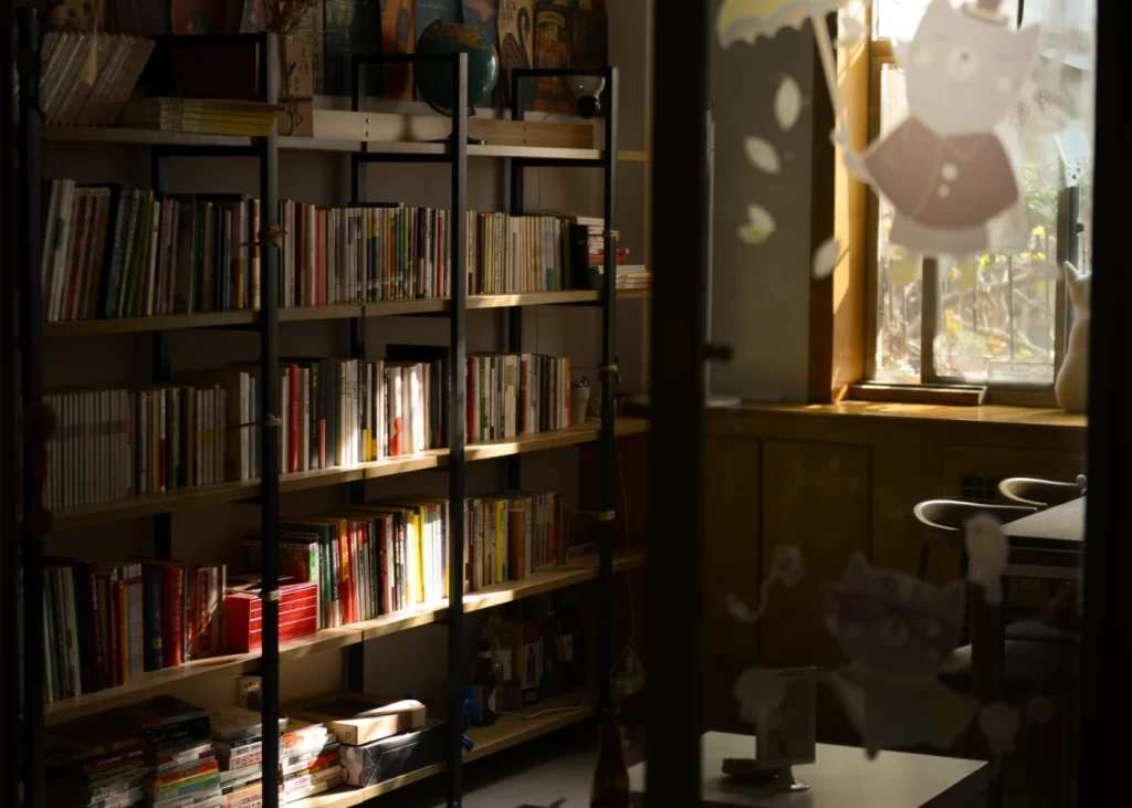کتابخانه ای که از سمت راست نور خورشید به دکور چوبی کتاب ها خورده است و در جلوی کتاب ها میز و صندلی و چراغ وجود دارد