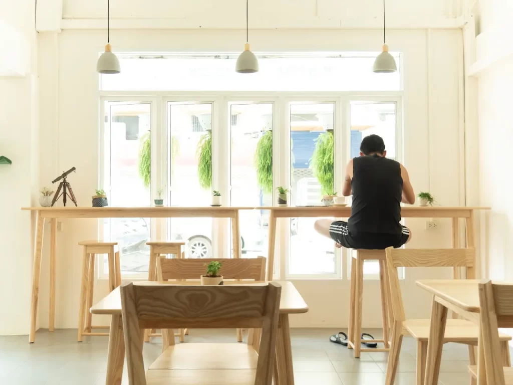 فردی که بر روی صندلی ها چوبی نشسته است و گیاهان در روی میز قرار دارد و پنجره هم نیز رو به روی فرد قرار دارد