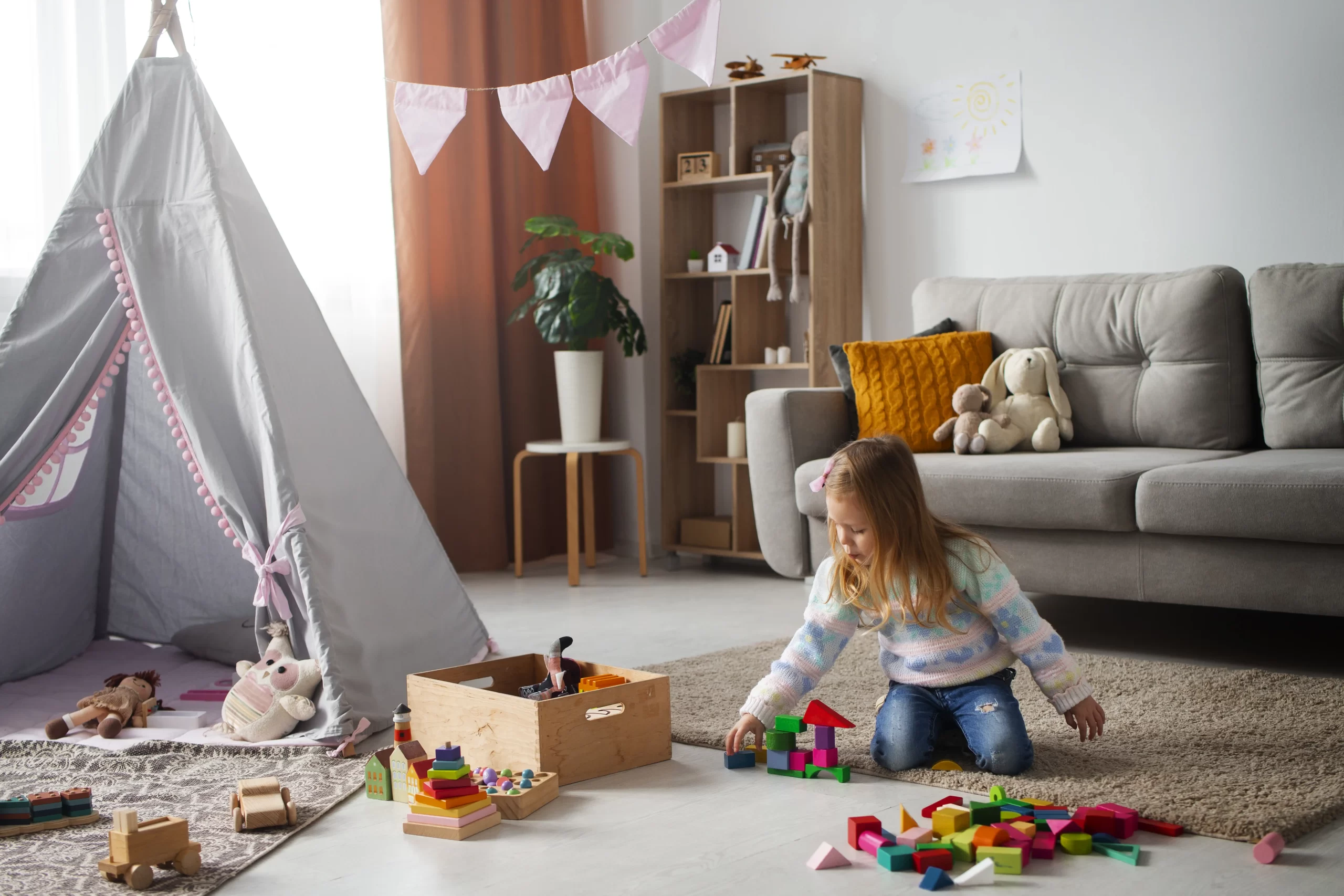دختری که در اتاقش است و وسایل بازیش بر روی زمین گذاشته است و مبل راحتی در پشتش قرار دارد و در کنار بچه چادر نیز قرار دارد