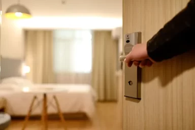 درب اتاق هتل در حال باز شدن است و تصویر محوی از یک تخت و میز در کنار آن نمایان است.